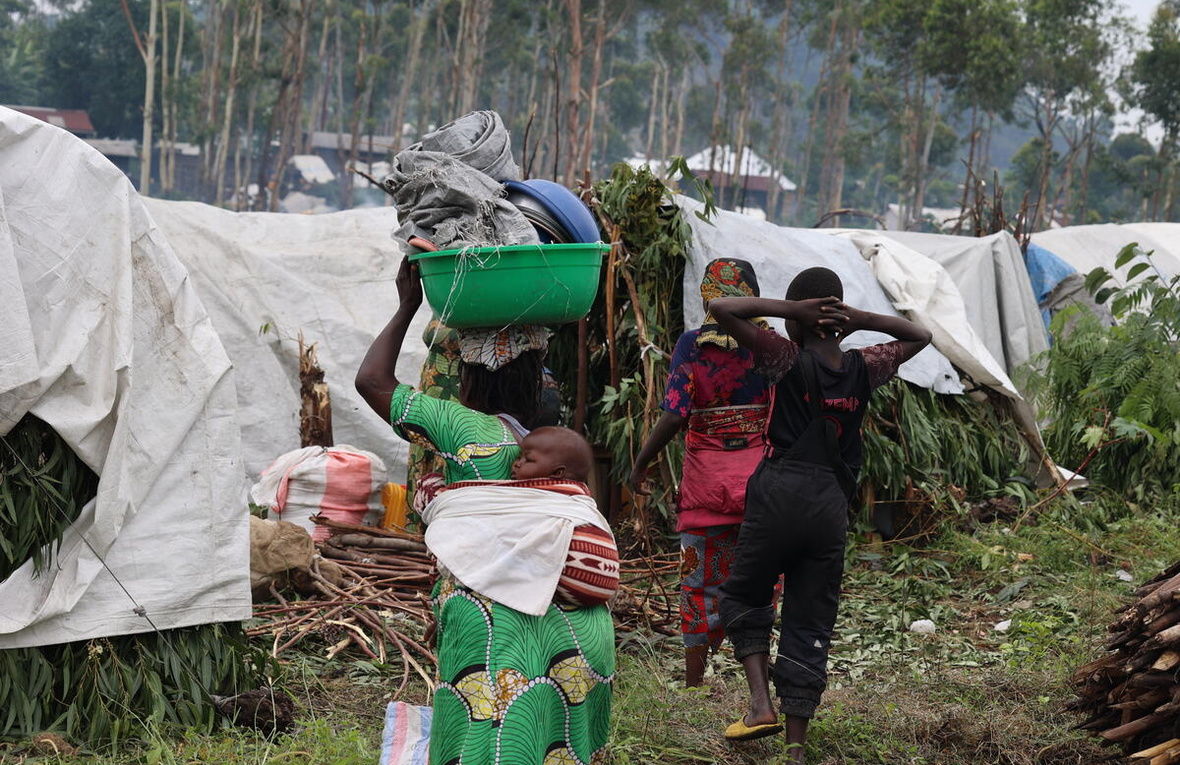 A woman walks towards makeshift shelters, her baby on her back and carrying a bucket of crockery and tarpaulin on her head. Two others walk alongside her.