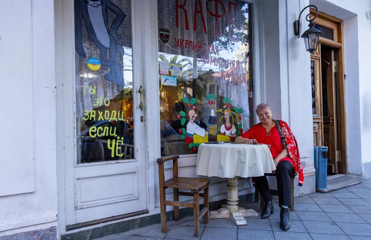 A smiling woman seated at a table in front of a café with colorful paintings on the windows and doors.