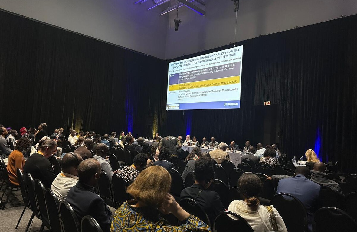 A large group of attendees at a conference look at a large screen with a PowerPoint presentation
