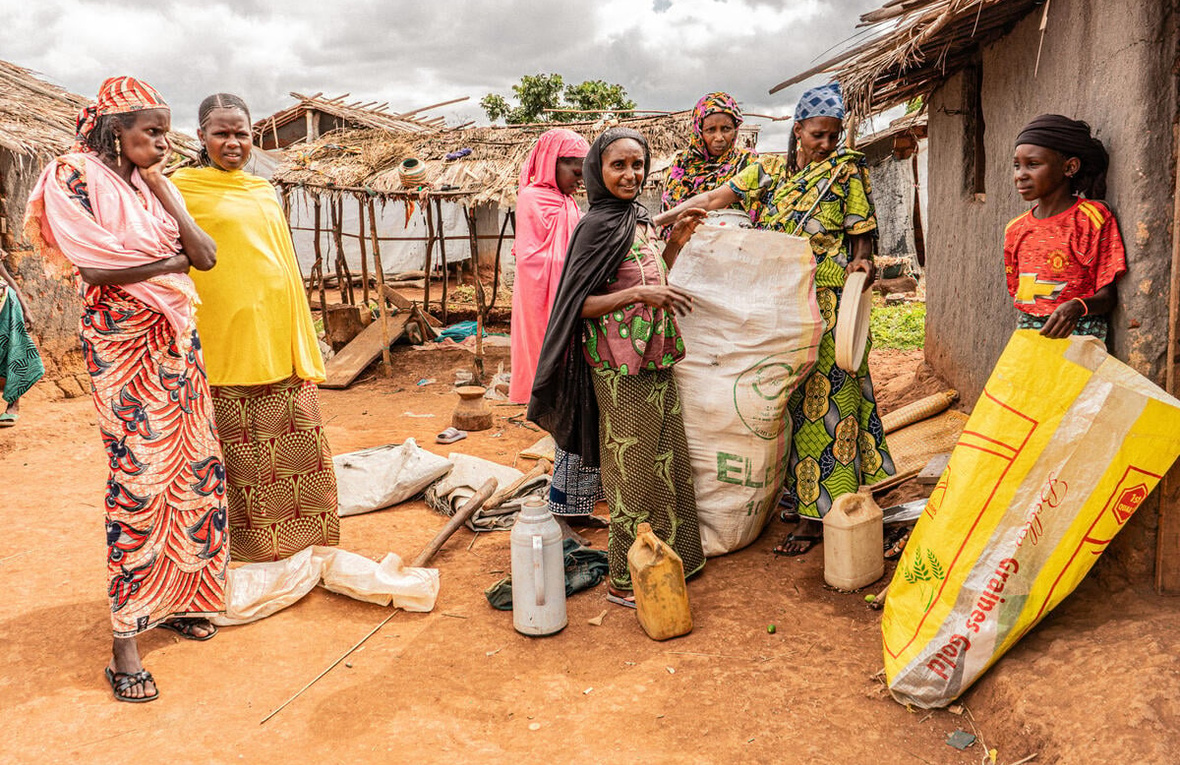 A group of women in colourful dresses stand outside a mud wall hut as they fill two large sacks with belongings