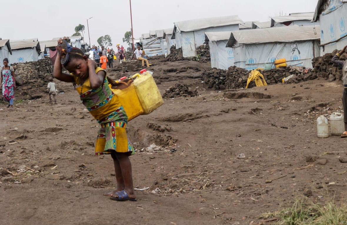 A young girl with two jerry cans on her back stands in front of a row of tents covered in tarpaulin.