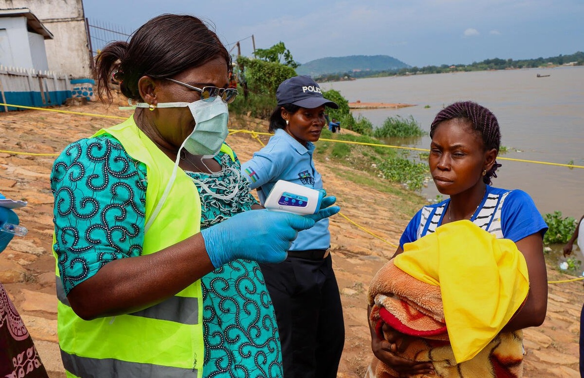A woman wearing a mask prepares to take another woman's temperature next to a river, while a police officer stands nearby.