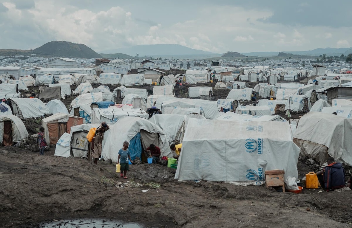 A young child walks across muddy ground in front of UNHCR tents with mountains in the background.
