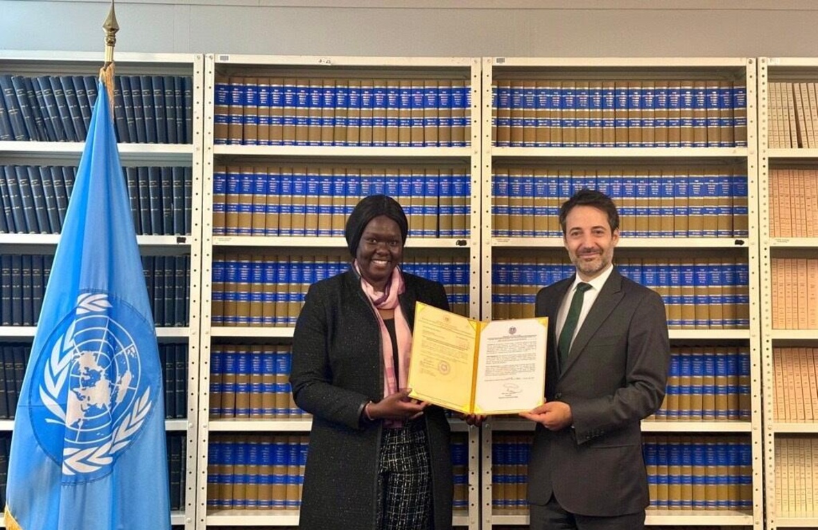 Cecilia Adeng and David Nanopoulos, both smiling proudly, stand in front of a large bookcase holding up a document folder, with a UN flag standing to the side.
