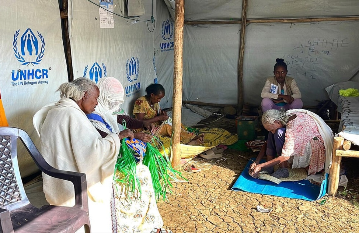 Several women sit inside a UNHCR tent and craft woven items