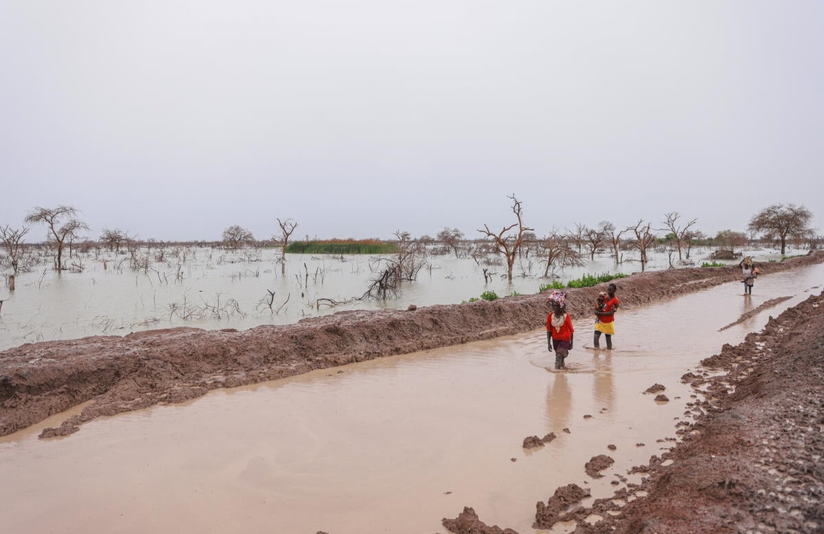 Adults carry children and belongings along a waterlogged road.