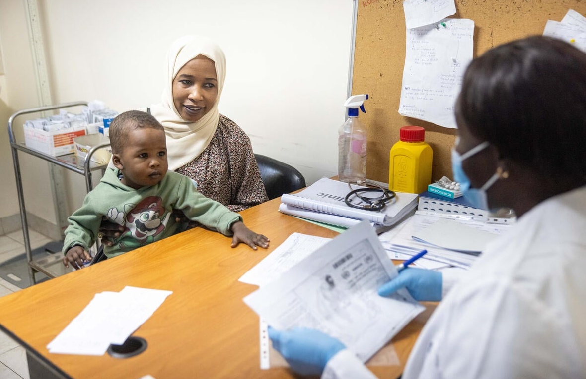 A woman is seated at a table with her young son, across from a medical staff member staff in a mask who is looking at paperwork