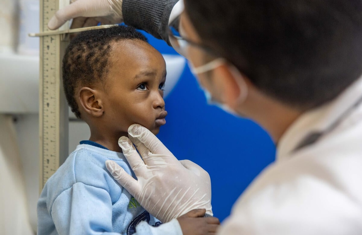 A young boy in a blue sweater stands against a height rule as a man wearing a surgical mask and gloves measures his height.