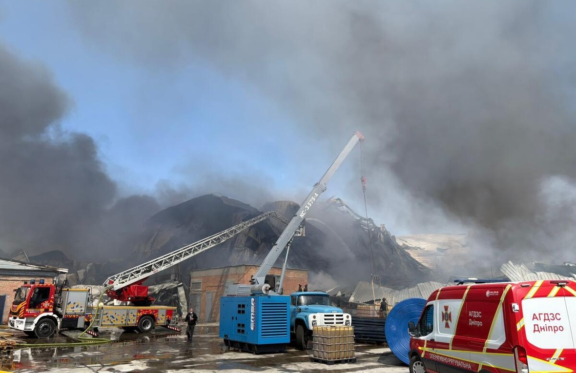 Black smoke surrounds a damaged warehouse, with emergency vehicles attending