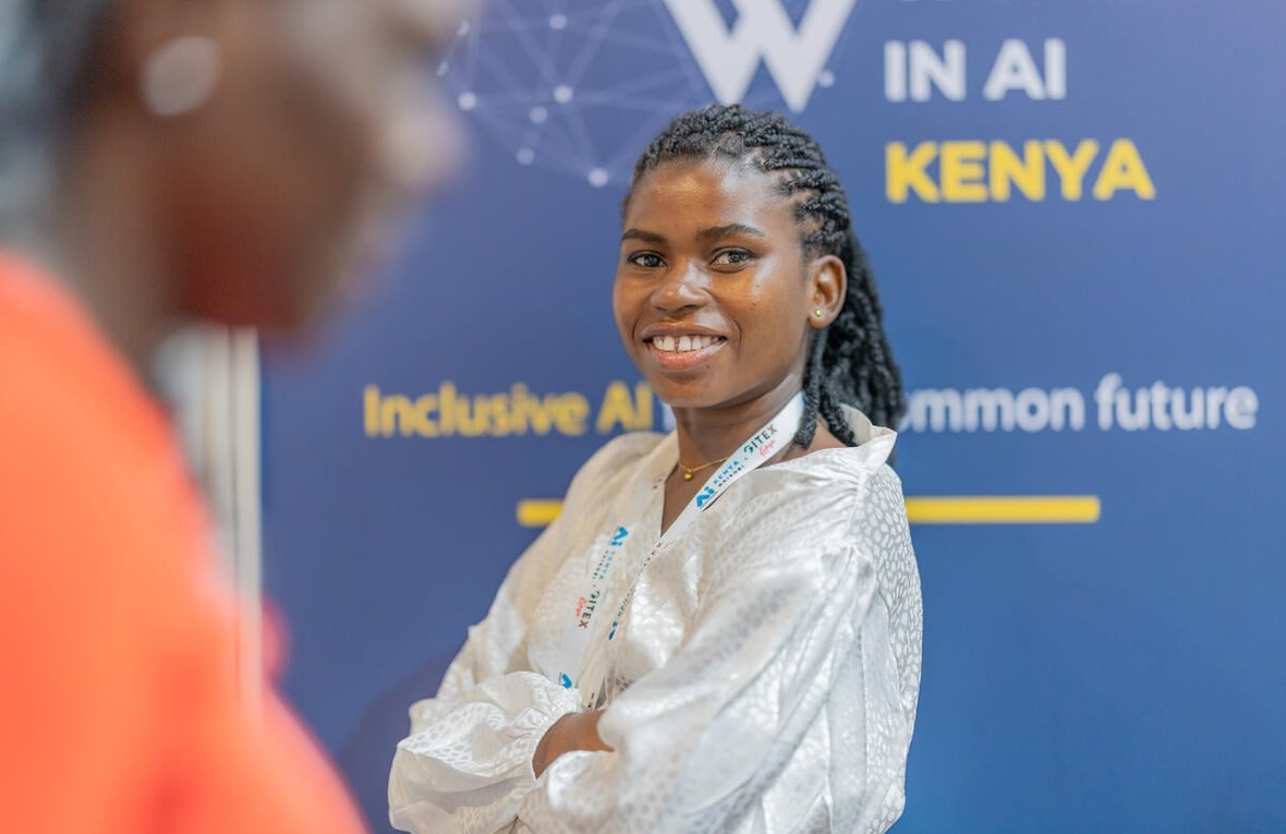 A smiling young woman with crossed arms stands in front of a conference poster for an event on women in AI in Kenya