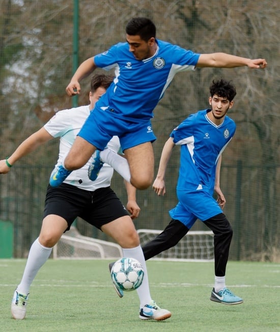 A football player jumps high in the air, the ball beneath him and other players behind him.