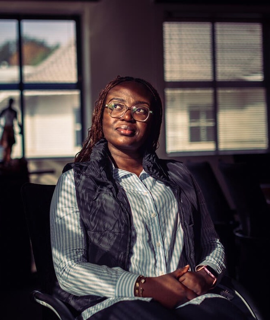 A woman sitting indoors in dappled light, with windows behind her, gazes into the distance