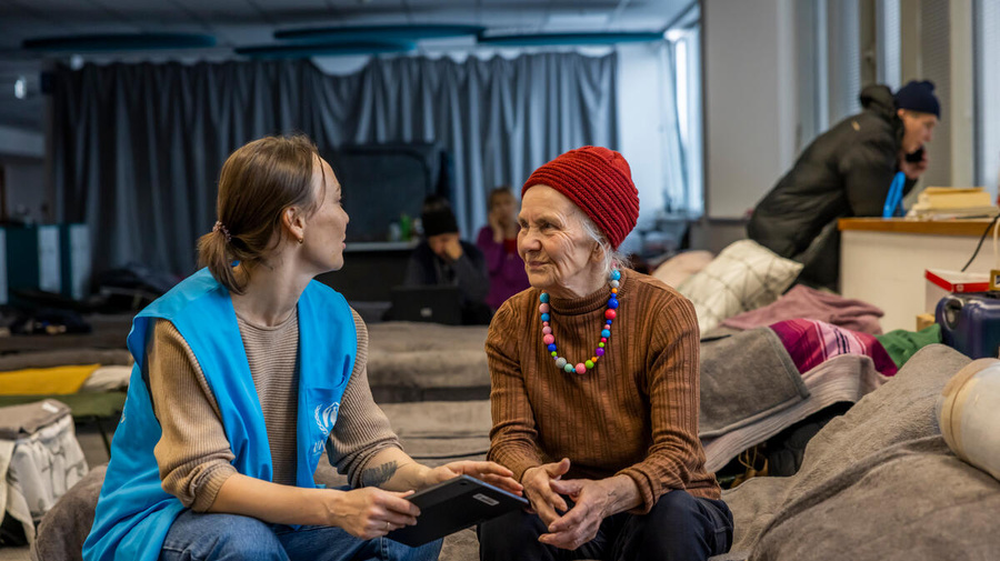 A UNHCR staff member speaks with a Ukrainian refugee inside a temporary shelter, a large hall filled with rows of beds.