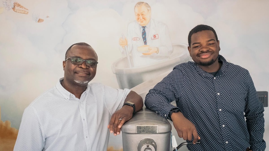 Two men stand either side of an industrial cake mixing machine in front of a mural-painted wall