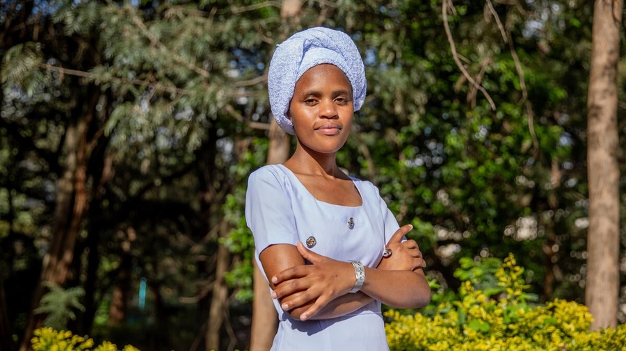 A woman in a blue dress and headscarf stands outside with her arms crossed and trees in the background