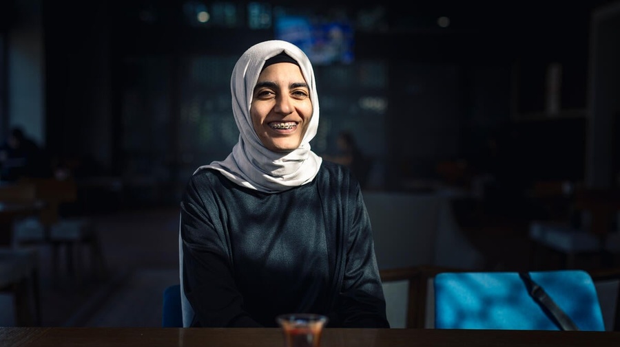 Aicha smiles widely at a table, a cup of tea in front of her