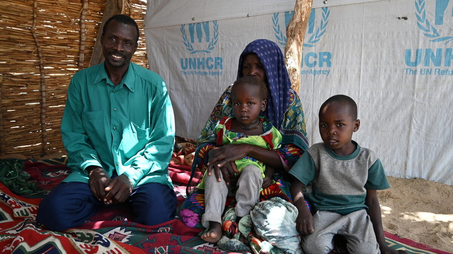 A couple sit on a carpet inside a woven reed shelter accompanied by two young children