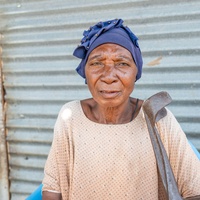 Older woman in blue headscarf standing outside her house