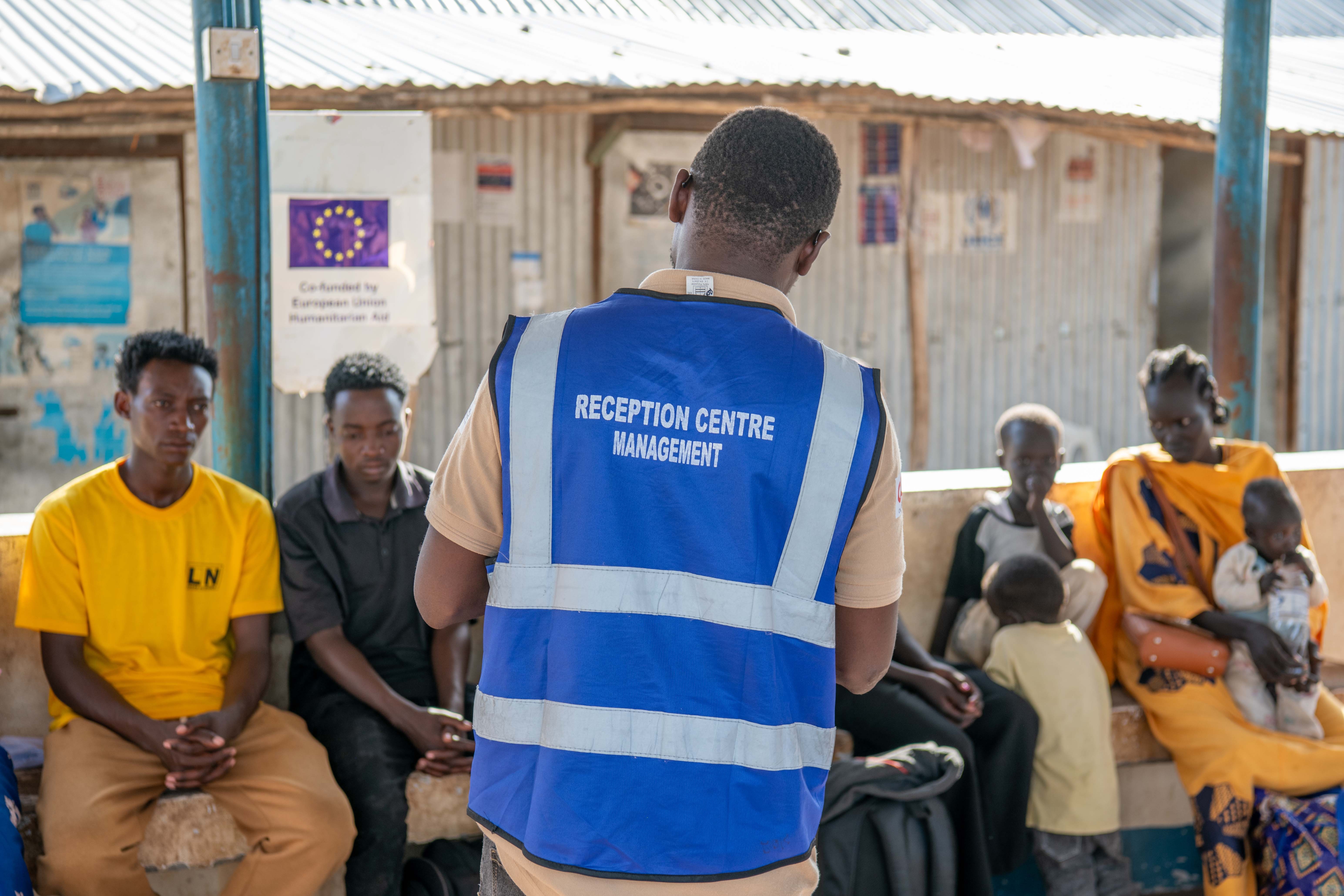 A staff member stands facing a group of newly arrived refugees seated at a reception centre in Kalobeyei.