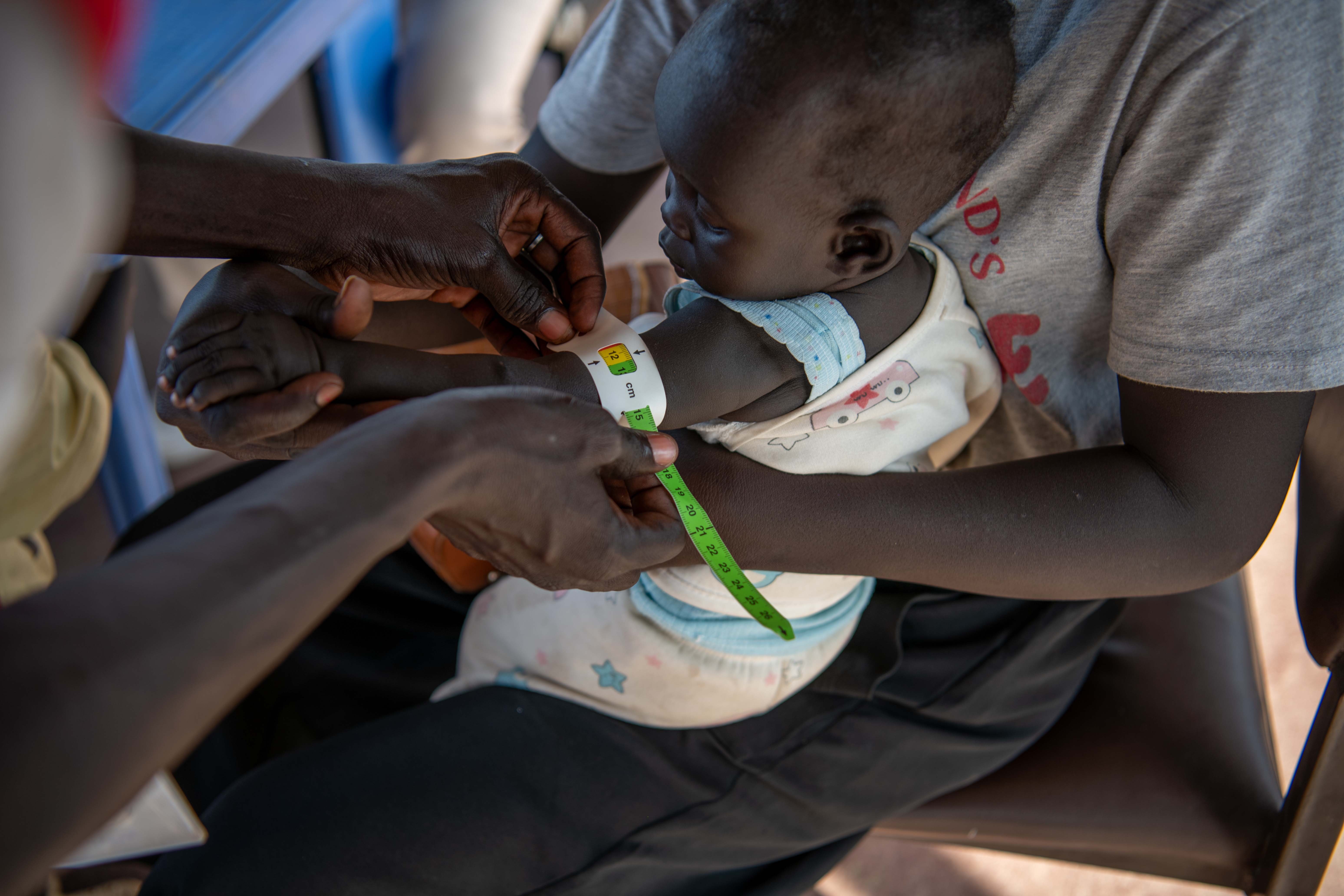 A baby sits on an adult&rsquo;s lap while a health worker measures the child&rsquo;s upper arm with a colour-coded tape.