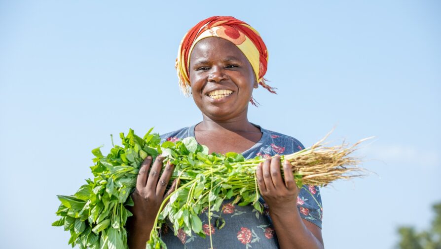 A smiling woman in a headscarf holds a bunch of green vegetables.