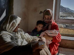 A mother cradles her baby in a bare room with her son and mother looking on