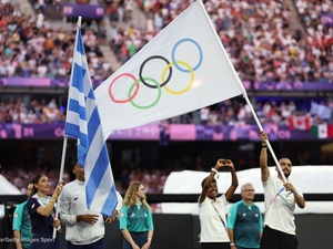 A man carries the Olympic flag in a packed stadium alongside a woman holding a Greek flag and others