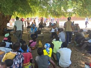 Un grand groupe de personnes se réunit sous un grand arbre. Des employés du HCR sont assis à de petits bureaux et prennent les enregistrements.