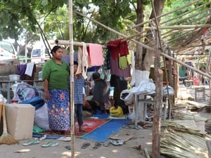 A woman stands under a tree with her arm around a young boy next to an unfinished bamboo structure and surrounded by belongings