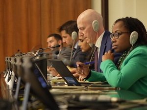 Close up of five speakers in a conference room, pictured with headphones on and laptops and microphones in front of them