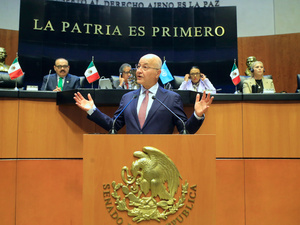 A man gestures while speaking behind a lectern with a row of people looking on from a raised platform behind