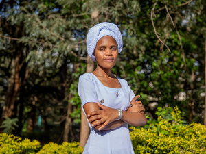A woman in a blue dress and headscarf stands outside with her arms crossed and trees in the background