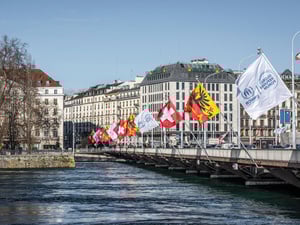 Flags fly along the length of a bridge over a wide river with buildings in the background
