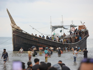 A group of people carrying belongings stand in the shallows alongside a wooden boat carrying refugees.