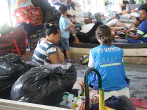 A UNHCR worker squats on the floor alongside refugees and their belongings in a temporary shelter