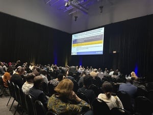 A large group of attendees at a conference look at a large screen with a PowerPoint presentation
