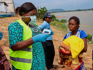 A woman wearing a mask prepares to take another woman's temperature next to a river, while a police officer stands nearby.
