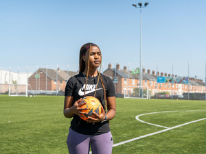 A young woman holds a football on a football pitch with rows of houses in the background