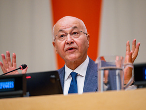 A speaker in glasses, a grey suit and blue tie addresses a briefing from behind a desk with a microphone and glass of water, both hands raised mid-gesture.