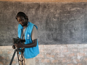 A man wearing a blue vest operates a camera in front of a blackboard with the writing: "I am Achouth. I was a refugee. And I am a filmmaker."