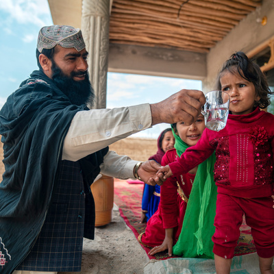 A man smiles as he holds a glass of water for a young girl to drink from.