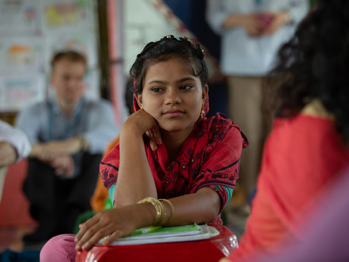 A girl dressed in red looks away from the camera while leaning on a small pile of books