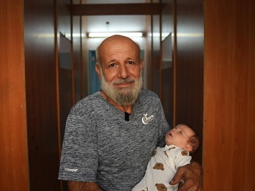 An older man with a beard holds a baby and looks into the camera.