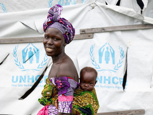 A smiling woman carries her baby boy on her back in front of a UNHCR tent.