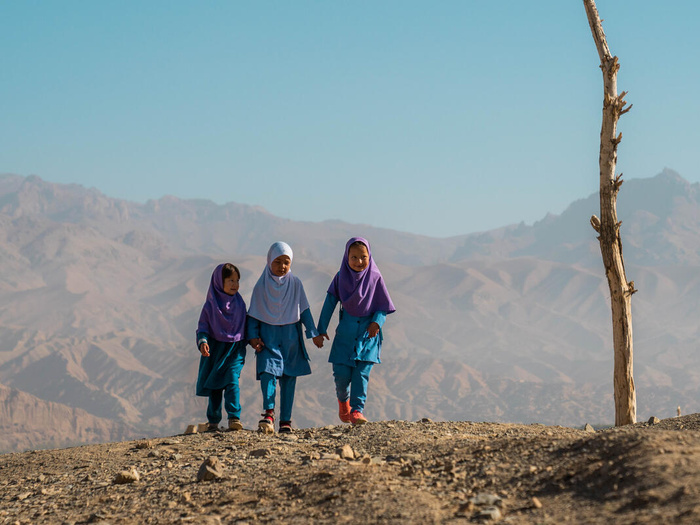 Three young female Afghan refugees walk hand in hand on arid ground, a mountain range behind them