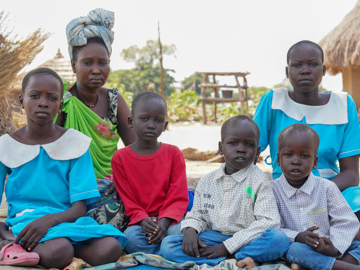 A family of 6 siblings sit together on a mat on the ground outside, some shelters with straw rooves behind them.