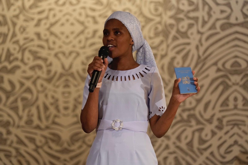 A woman on stage in front of a patterned backdrop speaks into a microphone while holding up a blue passport