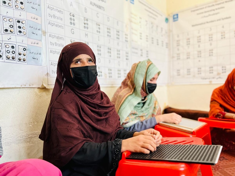 Helai Noori sits on the floor of a classroom with other women, reading in Braille from a wooden tablet.