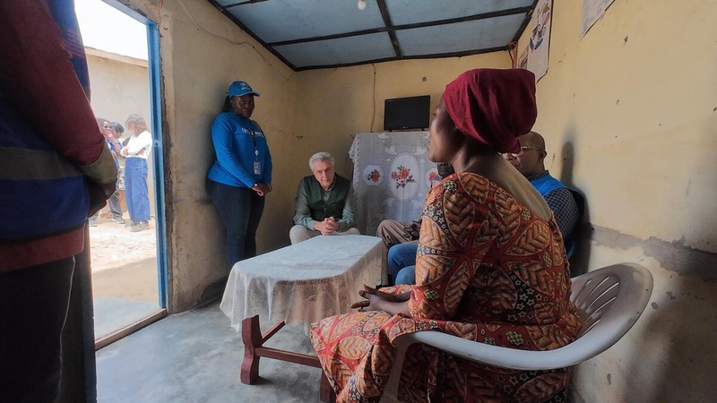 Une femme assise sur une chaise en plastique parle à des personnes assises et debout autour d'une table à l'intérieur d'un refuge.