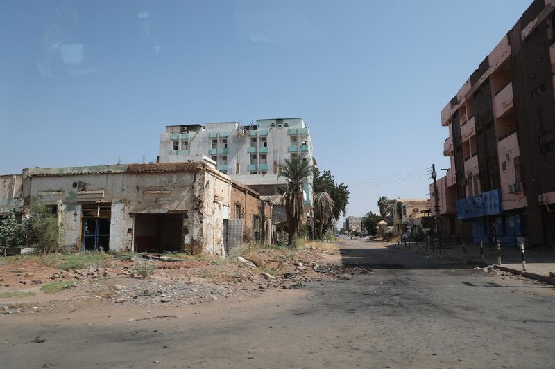 An empty street lined with palm trees, destroyed buildings and rubble.